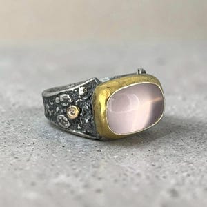 May include: A ring featuring a smooth, pale pink oval gemstone set in a gold bezel. The band is made of dark, oxidized silver with a textured, cratered surface and includes a small, round, flush-set gold accent near the main stone setting.