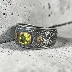 May include: A wide band ring featuring a textured, oxidized dark silver finish. The center is set with a square, faceted lime green gemstone in a gold-toned bezel, flanked by two small, circular gold-toned accents on the band's surface.