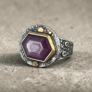 May include: A ring featuring a hexagonal purple gemstone set in a gold bezel. The ring band and surrounding frame are made of oxidized silver with a textured, pitted surface. Two small gold accents are embedded in the silver frame above and below.
