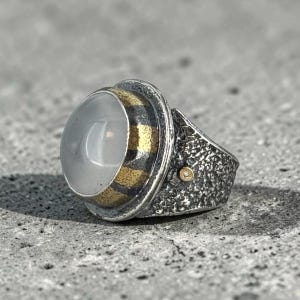 May include: A ring featuring a smooth, translucent, round cabochon stone set in a bezel with alternating oxidized silver and gold-toned stripes. The band has a heavily textured, dark, rugged surface and includes a small, circular gold-toned accent.