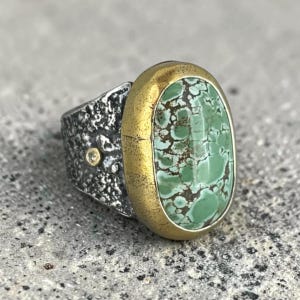 May include: A ring featuring an oval green turquoise stone with dark matrix patterns, set in a smooth gold bezel. The band is made of dark, textured oxidized silver and includes a small, round, clear gemstone accent embedded on the side.
