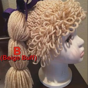 May include: A beige knitted hat with a curly hair design and a long ponytail made of yarn. The hat has a purple ribbon bow. The text "B (Beige Buff)" is visible on the hat.