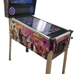May include: A Batman '66 pinball machine with a screen displaying gameplay. The cabinet features comic-style graphics of Batman characters, including Catwoman and the Joker, with the words "BANG" and "OUCH!" on the sides. The machine has silver legs.