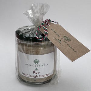 May include: A jar of rye sourdough starter, wrapped in cellophane and tied with red, green, and white twine. A brown tag with the words "Green Cottages Kitchen" is attached. The jar has a black lid.