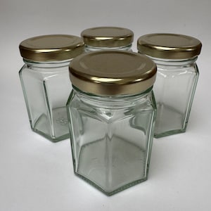 May include: Four clear glass hexagonal jars with gold lids. The jars are empty and have a smooth, polished finish.
