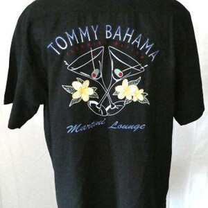 May include: Black short-sleeved shirt with embroidered design. The back features the text "TOMMY BAHAMA" above a martini glass graphic with yellow flowers and the words "Martini Lounge".