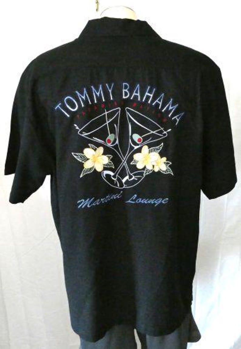 May include: Black short-sleeved shirt with embroidered design. The back features the text "TOMMY BAHAMA" above a martini glass graphic with yellow flowers and the words "Martini Lounge".