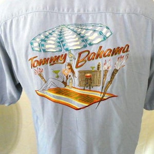 May include: A light blue button-down shirt with a Tommy Bahama logo and a tropical scene embroidered on the back. The scene features a woman relaxing under an umbrella with a cocktail.