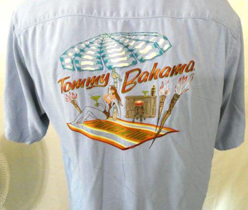 May include: A light blue button-down shirt with a Tommy Bahama logo and a tropical scene embroidered on the back. The scene features a woman relaxing under an umbrella with a cocktail.