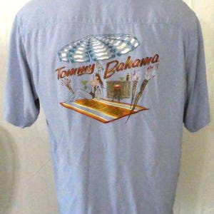 May include: A light blue button-down shirt with a Tommy Bahama logo and a tropical scene printed on the back. The scene features a woman in a bikini under a beach umbrella with palm trees and a bar in the background.