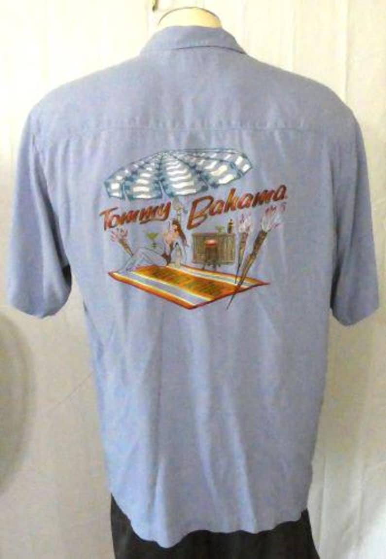 May include: A light blue button-down shirt with a Tommy Bahama logo and a tropical scene printed on the back. The scene features a woman in a bikini under a beach umbrella with palm trees and a bar in the background.