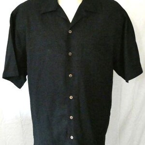 May include: Black short-sleeved button-down shirt with a classic collar. The shirt has a subtle textured pattern and is buttoned up the front with six buttons. The shirt is displayed on a mannequin.
