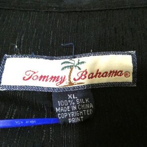 May include: Close-up of a black silk shirt tag. The tag reads "Tommy Bahama" in red script with a palm tree graphic. Below the main tag is a smaller tag with text including "XL" and "100% Silk."
