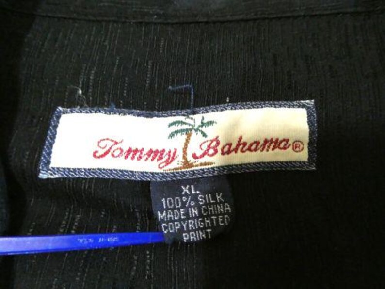 May include: Close-up of a black silk shirt tag. The tag reads "Tommy Bahama" in red script with a palm tree graphic. Below the main tag is a smaller tag with text including "XL" and "100% Silk."