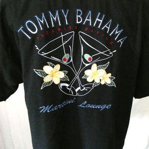 May include: Black short-sleeved shirt with the text "TOMMY BAHAMA PARADISE NATION" in white and blue. The shirt features a graphic of two martini glasses with olives, and yellow flowers with green leaves. The text "Martini Lounge" is also visible.