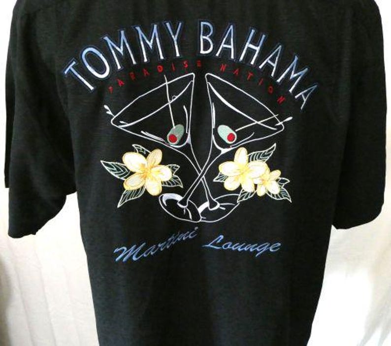 May include: Black short-sleeved shirt with the text "TOMMY BAHAMA PARADISE NATION" in white and blue. The shirt features a graphic of two martini glasses with olives, and yellow flowers with green leaves. The text "Martini Lounge" is also visible.