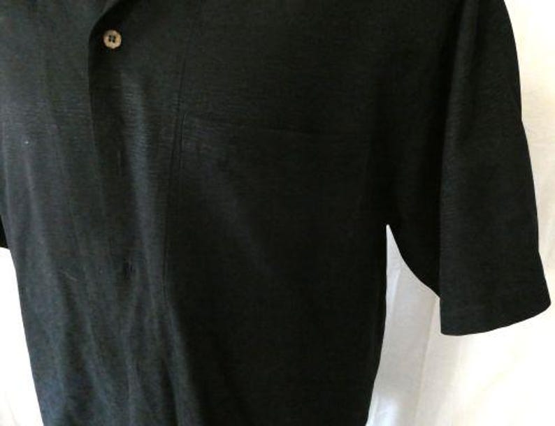 May include: Black short-sleeved shirt with a button-down collar and a single chest pocket. The shirt has a textured appearance and a button closure. The fabric appears to be a lightweight material.