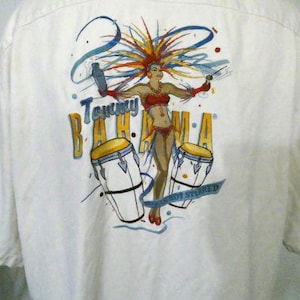Pode incluir: Camisa branca de botões com uma ilustração colorida de uma mulher com um chapéu de penas e um biquíni, segurando um coquetel. O texto "Tommy Bahama" e "Not Stirred" está impresso na camisa.