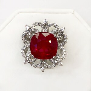 May include: A red gemstone ring with a cushion-cut ruby surrounded by a halo of smaller diamonds. The ring setting is a delicate, intricate design with a floral motif.