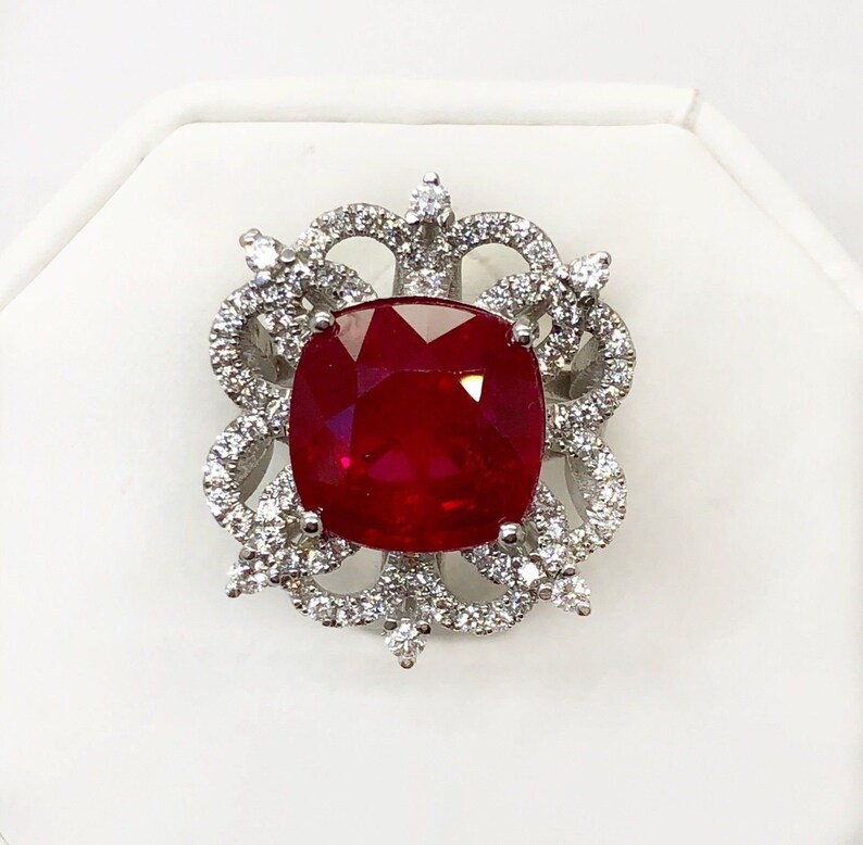 May include: A red gemstone ring with a cushion-cut ruby surrounded by a halo of smaller diamonds. The ring setting is a delicate, intricate design with a floral motif.