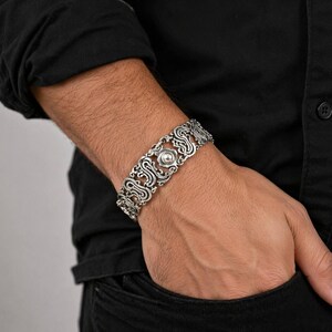 K&ouml;nnte beinhalten: Silberfarbenes Armband mit einem verzierten, durchbrochenen Design. Das Armband hat ein zentrales, kreisf&ouml;rmiges Element und sich wiederholende Scrollmuster. Der Schmuck wird am Handgelenk getragen und zeigt seine komplizierten Details.