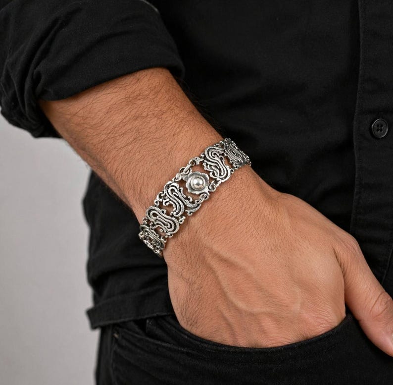 K&ouml;nnte beinhalten: Silberfarbenes Armband mit einem verzierten, durchbrochenen Design. Das Armband hat ein zentrales, kreisf&ouml;rmiges Element und sich wiederholende Scrollmuster. Der Schmuck wird am Handgelenk getragen und zeigt seine komplizierten Details.