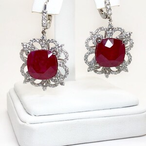 May include: A pair of dangle earrings with a large red gemstone surrounded by a halo of smaller white gemstones. The earrings are made of silver and have a hook closure.