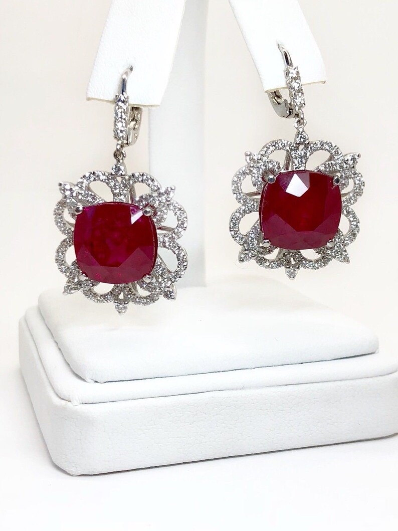 May include: A pair of dangle earrings with a large red gemstone surrounded by a halo of smaller white gemstones. The earrings are made of silver and have a hook closure.