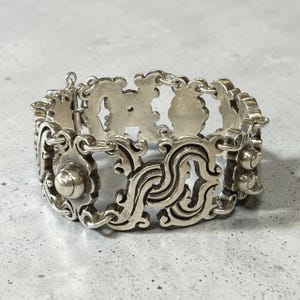 K&ouml;nnte beinhalten: Ein silberfarbenes Armband mit einem kunstvollen, durchbrochenen Design. Das Armband besteht aus miteinander verbundenen, dekorativen Gliedern mit wirbelnden Mustern und abgerundeten Akzenten. Der Verschluss ist sichtbar, und der Stil deutet auf eine Vintage- oder Antik-&Auml;sthetik hin. Ca. 18 cm lang.