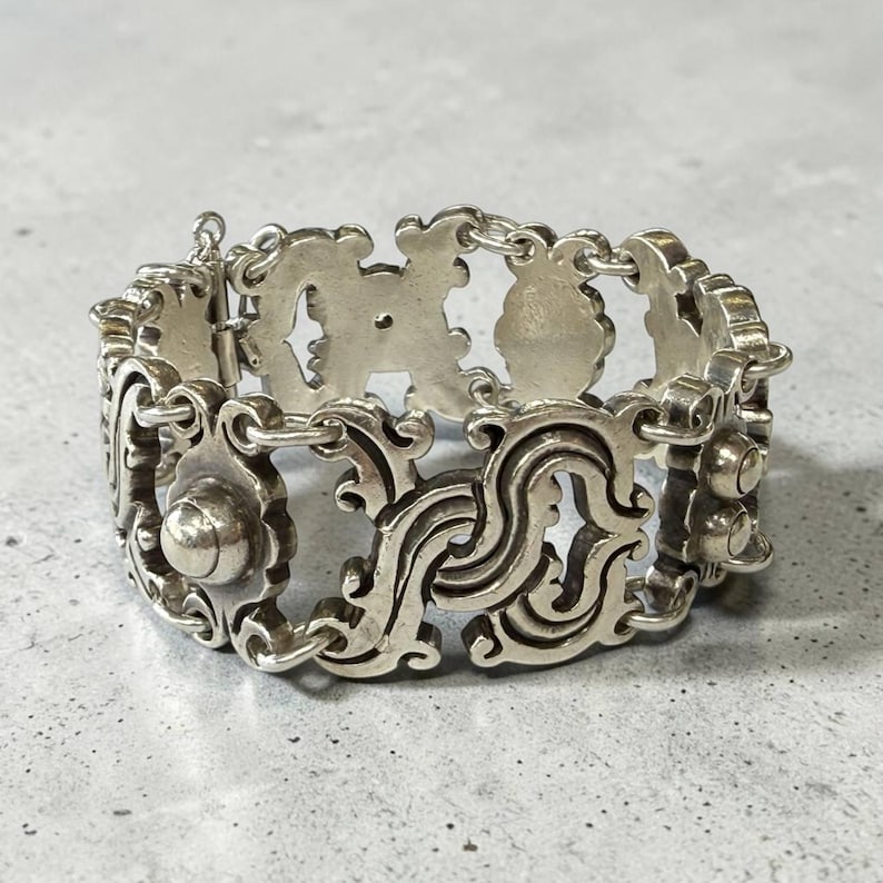 K&ouml;nnte beinhalten: Ein silberfarbenes Armband mit einem kunstvollen, durchbrochenen Design. Das Armband besteht aus miteinander verbundenen, dekorativen Gliedern mit wirbelnden Mustern und abgerundeten Akzenten. Der Verschluss ist sichtbar, und der Stil deutet auf eine Vintage- oder Antik-&Auml;sthetik hin. Ca. 18 cm lang.