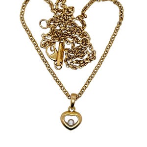 Puede incluir: Un collar dorado con un colgante en forma de corazón. El colgante tiene una pequeña gema transparente en el centro. La cadena está dispuesta en forma de corazón. El collar se muestra sobre un fondo blanco.