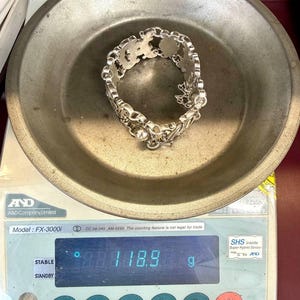 K&ouml;nnte beinhalten: Ein silbernes Armband mit einem verzierten Design liegt auf einer Metallwaage. Das Armband besteht aus einer Reihe von miteinander verbundenen dekorativen Gliedern. Die Waage zeigt ein Gewicht von 118,9 Gramm an. Die Waage befindet sich auf einer wei&szlig;en und roten Oberfl&auml;che.