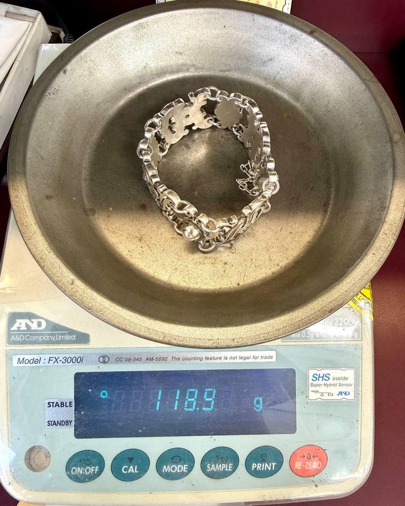 K&ouml;nnte beinhalten: Ein silbernes Armband mit einem verzierten Design liegt auf einer Metallwaage. Das Armband besteht aus einer Reihe von miteinander verbundenen dekorativen Gliedern. Die Waage zeigt ein Gewicht von 118,9 Gramm an. Die Waage befindet sich auf einer wei&szlig;en und roten Oberfl&auml;che.