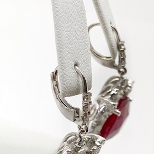 May include: A pair of dangle earrings with a large red gemstone surrounded by smaller white gemstones. The earrings have a silver hook closure.