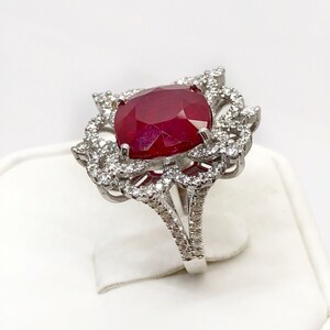 May include: A silver ring with a large, red ruby gemstone surrounded by smaller diamonds. The ring has a delicate, floral design.