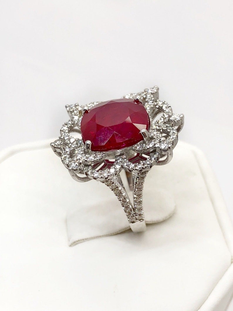 May include: A silver ring with a large, red ruby gemstone surrounded by smaller diamonds. The ring has a delicate, floral design.