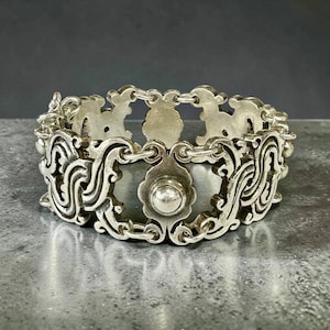 K&ouml;nnte beinhalten: Ein silberfarbenes Armband mit einem verzierten, durchbrochenen Design. Das Armband weist sich wiederholende Blumen- und Scrollmotive auf, die durch kleine Kettenglieder verbunden sind. Auf jeder dekorativen Platte befindet sich ein zentrales, kreisf&ouml;rmiges Element.