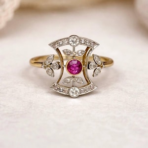 May include: A vintage-style ring with a gold band and a decorative setting. The setting features a central, round, bright pink gemstone, surrounded by smaller clear stones and leaf-like accents. The ring's design is symmetrical and ornate.