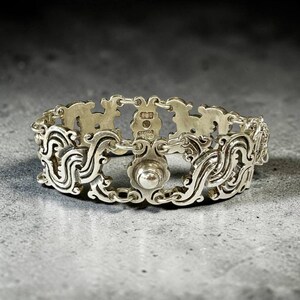 K&ouml;nnte beinhalten: Ein silberfarbenes Armband mit einem verzierten, durchbrochenen Design. Das Armband weist geschwungene Muster und ein zentrales, kreisf&ouml;rmiges Element auf. Das Metall hat eine polierte Oberfl&auml;che, die das Licht reflektiert. Das Armband ist wahrscheinlich aus Sterlingsilber oder einem &auml;hnlichen Metall gefertigt.