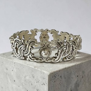 K&ouml;nnte beinhalten: Ein silberfarbenes Armband mit einem kunstvollen, geschwungenen Design. Das Armband besteht aus miteinander verbundenen Gliedern mit einem zentralen, kreisf&ouml;rmigen Verschluss. Die detaillierten Elemente umfassen Wirbelmuster und dekorative Elemente.