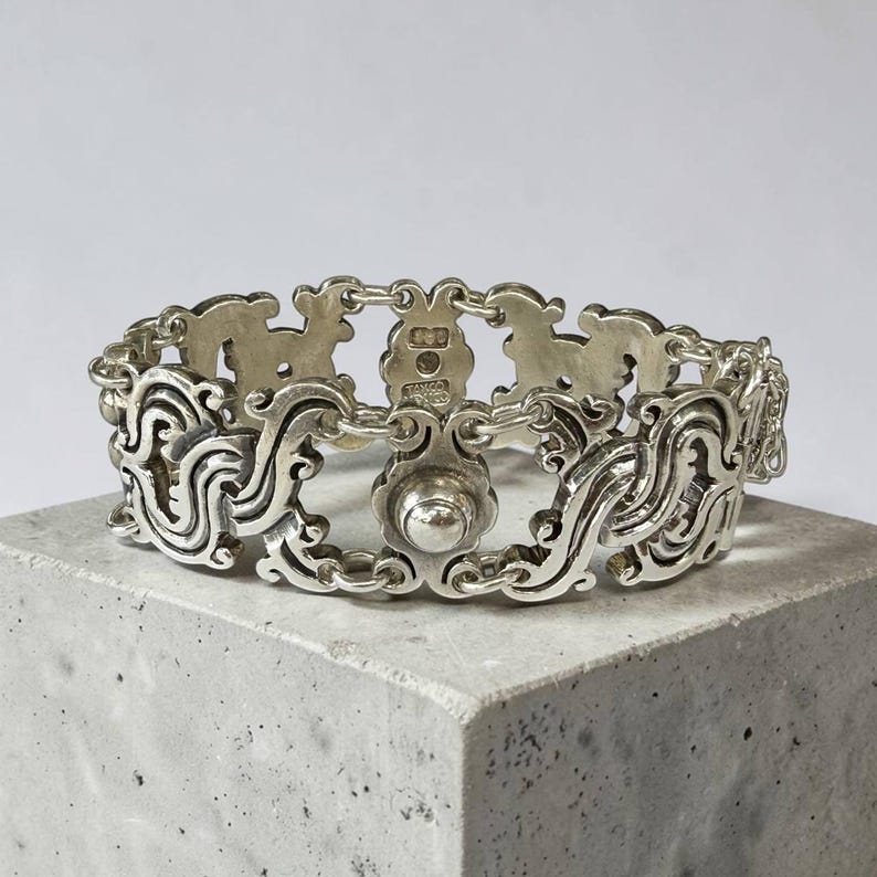 K&ouml;nnte beinhalten: Ein silberfarbenes Armband mit einem kunstvollen, geschwungenen Design. Das Armband besteht aus miteinander verbundenen Gliedern mit einem zentralen, kreisf&ouml;rmigen Verschluss. Die detaillierten Elemente umfassen Wirbelmuster und dekorative Elemente.
