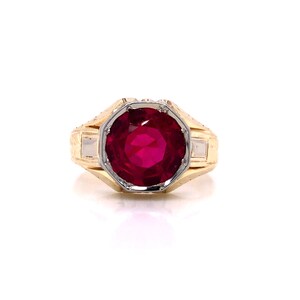 May include: A gold ring featuring a large, red gemstone set in a white metal bezel. The ring has a textured band with an ornate design.