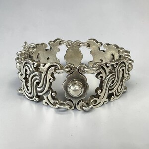 K&ouml;nnte beinhalten: Ein silberfarbenes Armband mit einem verzierten, durchbrochenen Design. Das Armband weist sich wiederholende Blumen- und Scrollmotive auf, die durch kleine Kettenglieder verbunden sind. Das zentrale Designelement ist ein bl&uuml;tenf&ouml;rmiges St&uuml;ck mit einem runden, erhabenen Mittelteil.