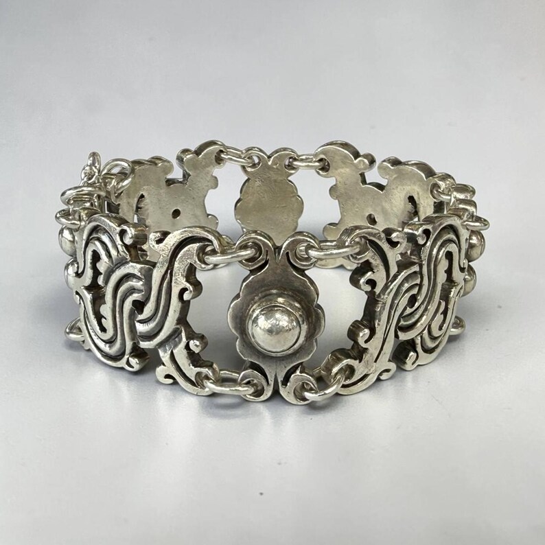 K&ouml;nnte beinhalten: Ein silberfarbenes Armband mit einem verzierten, durchbrochenen Design. Das Armband weist sich wiederholende Blumen- und Scrollmotive auf, die durch kleine Kettenglieder verbunden sind. Das zentrale Designelement ist ein bl&uuml;tenf&ouml;rmiges St&uuml;ck mit einem runden, erhabenen Mittelteil.