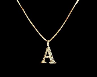 Puede incluir: Un collar de cadena de oro con un colgante en forma de la letra A. El colgante está hecho de oro y tiene una superficie texturizada.