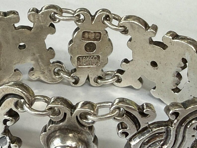 K&ouml;nnte beinhalten: Nahaufnahme eines Silberarmbands mit verzierten, stilisierten floralen und scrollartigen Gliedern. Das Armband ist durch kleine, runde Kettenglieder verbunden. Der Verschluss ist mit "925" und "TAXCO MEXICO" gestempelt.