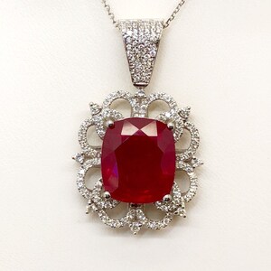 May include: A silver pendant necklace with a large, red, cushion-cut ruby surrounded by smaller diamonds. The pendant is set in a delicate, ornate design.