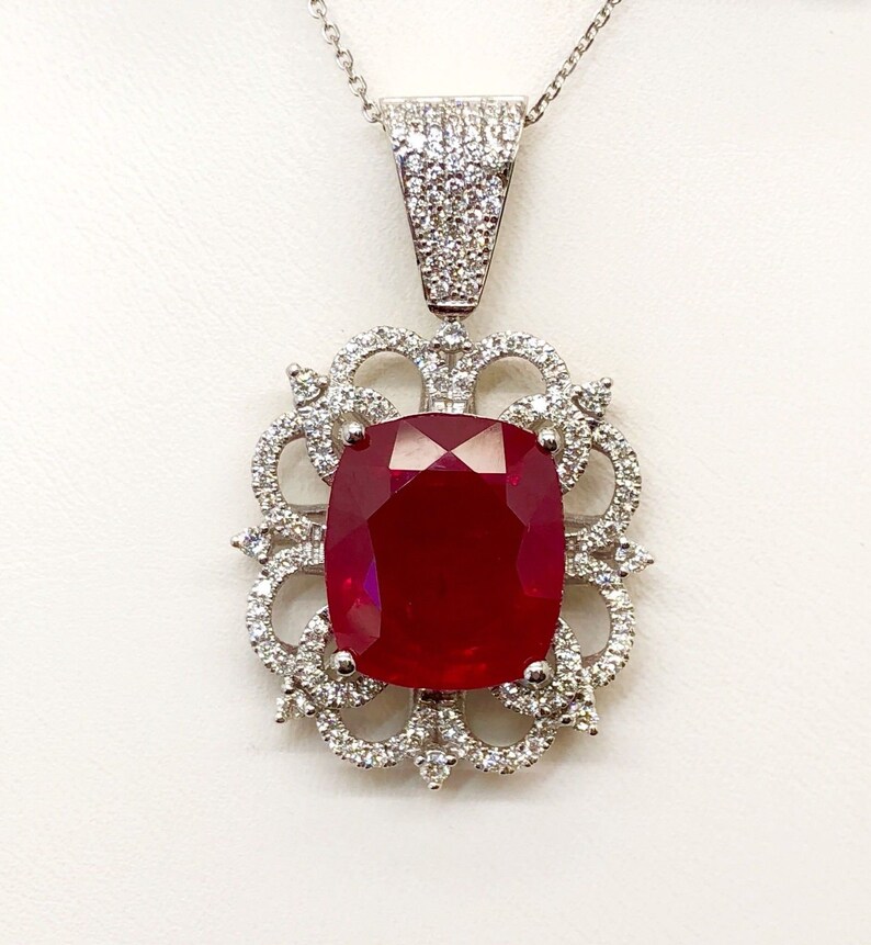 May include: A silver pendant necklace with a large, red, cushion-cut ruby surrounded by smaller diamonds. The pendant is set in a delicate, ornate design.