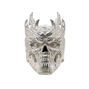 May include: A silver skull ring with a detailed design of flames and horns. The ring is a statement piece with a gothic and edgy aesthetic.