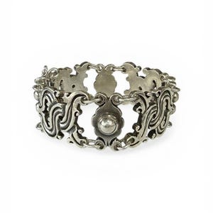 K&ouml;nnte beinhalten: Ein silberfarbenes Armband mit einem verzierten, durchbrochenen Design. Das Armband weist sich wiederholende Blumen- und Scrollmuster auf, die durch kleine Kettenglieder verbunden sind. Ein zentrales bl&uuml;tenf&ouml;rmiges Element mit erhabenem Mittelpunkt f&uuml;gt Details hinzu.